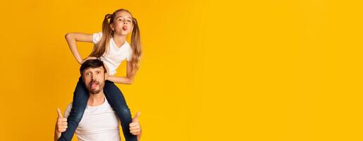 A father gives his daughter a fun ride on his shoulders while both make playful faces, capturing a joyful moment of their bond and laughter. Vibrant yellow backdrop enhances the cheerful atmosphere. photo