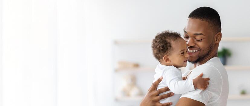 niño cuidado. retrato de contento africano americano padre con infantil bebé en brazos en pie cerca ventana a hogar, joven negro papá gasto hora con adorable niñito niño, panorama con Copiar espacio foto