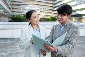 Asian man and woman are outside in a contemporary urban environment, discussing and reviewing documents together. They appear to be collaborating on a project or presentation photo