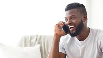 African American young man, wearing a white t-shirt and sitting on a couch at home, is talking on the phone while smiling broadly. The man has a short haircut and a full beard, copy space photo