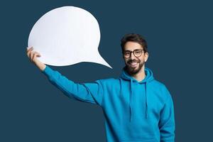 A man with glasses is smiling while holding a large blank speech bubble in the air. He is dressed in a blue hoodie, creating a playful, engaging atmosphere suitable for various creative uses. photo