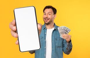 A smiling man displays a blank smartphone in one hand while holding a stack of cash in the other. The vibrant yellow background enhances the cheerful atmosphere of the moment. photo