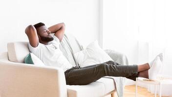Black man sits on a white couch with his arms behind his head, relaxing and enjoying a moment of peace. His feet are up on a small table beside the couch, and he appears to be taking a break photo
