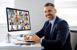 A businessman in a suit sits at a desk in an office, looking at the camera while participating in a conference meeting on a computer monitor. He is smiling and holding a pen in his hand photo