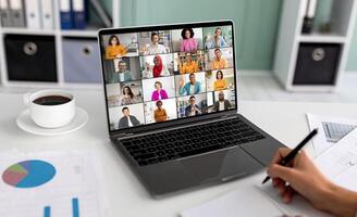 A laptop screen displays a conference meeting with a grid of multiple participants, all of whom are smiling and waving. Computer is positioned on a white desk in front of a person who is writing photo