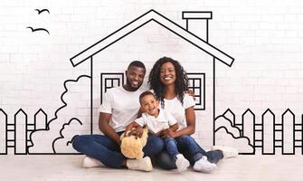 Happy black family of three father mother and little son sitting on floor against illustrated house of their dreams over white wall background photo
