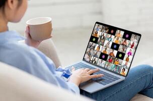 A woman is sitting on a couch, holding a cup of coffee in one hand and using a laptop with a conferencing app open. The screen shows numerous participants in the call. photo