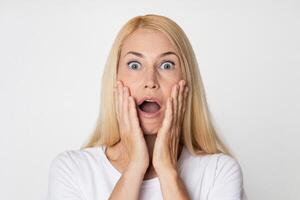 This image shows a woman with blonde hair looking shocked, with her mouth open and her hands covering her cheeks. She is dressed in a white t-shirt and is looking directly at the camera. photo