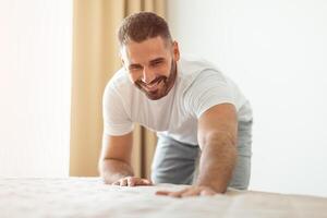 A man in a white shirt and grey pants smiles while making his bed in his bedroom. He is kneeling on the bed, smoothing out the sheets with one hand. Sunlight streams through a window behind him. photo