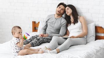 A young couple sits exhausted on a bed, while their baby plays with a toy on the white sheets. The father looks tired and leans back, while the mother seems to be nodding off. photo