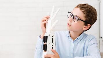 A young boy wearing glasses examines a robotic hand in a classroom setting. He appears to be intrigued and fascinated by the technology, demonstrating the growing interest in robotics photo