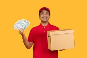 A cheerful delivery guy dressed in a red uniform proudly displays a fan of cash in one hand while holding a cardboard package in the other. The bright yellow background photo