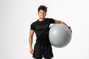 A young athlete stands confidently in a studio gym setting, holding a large exercise ball with a smile on his face. He is dressed in a black athletic outfit photo