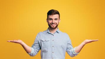 Smiling millennial man holding empty palms up as scales, making choice, copy space, yellow studio background photo
