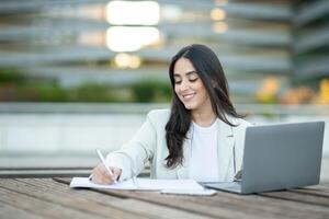 A woman seated at a wooden table enjoys writing notes while using a laptop in an urban setting. photo