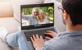 A man is sitting on a couch and chatting with an older couple on his laptop. He is smiling and appears to be engaged in conversation. photo