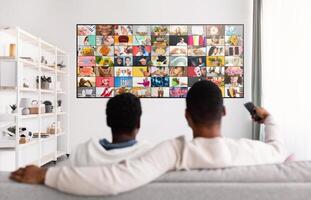 Two men sit on a white couch facing a large flat-screen television with a grid of various images on the screen. One man is holding a remote control in his right hand. photo