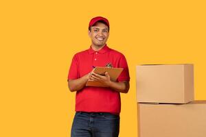 A cheerful delivery guy in a red uniform holds a clipboard while standing beside cardboard boxes. The bright yellow background highlights his friendly demeanor as he prepares for a package delivery. photo