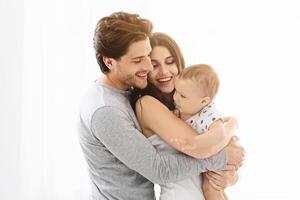 This image shows a happy family of three, a man, a woman, and their baby, standing close together in a white interior setting. The man and woman are smiling at the baby photo