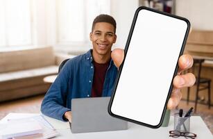 African American young man sits at a desk in a well-lit room, working on a laptop. He holds up a smartphone, smiling cheerfully, ready to share information or a message. photo
