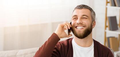 A man sitting comfortably on a couch, engaged in a phone conversation. He is holding a cell phone to his ear, gesturing while speaking. The background is a typical living room setting. photo