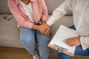 A therapist sits beside their patient on a couch and holds their hand, offering comfort and support. The therapist is taking notes on a clipboard, while the patient appears to be listening intently. photo