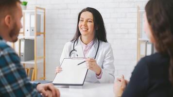 Woman doctor wearing a white coat is sitting at a table and discussing medical information with a couple. She is holding a clipboard and pointing at it with a pen. photo
