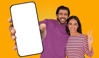 A young man and woman are smiling and posing for a photo in front of a bright yellow background. The man is holding a smartphone with a blank screen, extending it towards the camera
