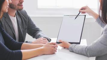 A businesswoman in a gray blazer is presenting a contract to a young couple in an office setting. She is holding a clipboard and pointing at the document with a pen. photo