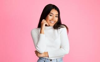 Asian young woman with long black hair is smiling and looking at the camera. She is wearing a white sweater and has her hand on her chin. The background is a solid pink color. photo