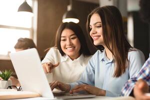 Two multiethnic women are seated at a desk in a well-lit library, working together on a project. One is pointing at a laptop screen, suggesting an idea or solution photo