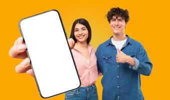 A young man and woman stand in front of a yellow background. The woman is holding a smartphone with a blank white screen towards the viewer, while the man gives a thumbs up. Both are smiling. photo