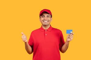 A cheerful delivery man dressed in a red uniform is giving a thumbs up gesture while holding a blue card. The background is a vibrant yellow, adding to the positive and energetic atmosphere photo