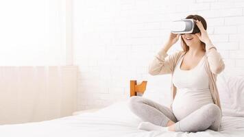 A pregnant woman is sitting on a bed, engrossed in using a virtual reality device. She appears focused and engaged with the technology, experiencing a virtual environment, copy space photo