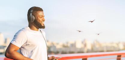 alegre africano hombre teniendo descanso mientras hacer ejercicio en el puente, escuchando a música. disfrutando ciudad vista, Copiar espacio foto