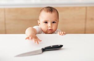 A curious baby stretches out its arm, reaching for a knife placed on a kitchen table under bright morning light. The child is focused and intrigued by the object within its grasp. photo