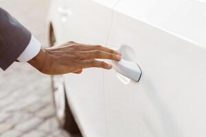 A closeup image of a hand reaching for the door handle of a white car. The hand is in the foreground, and the car door is in the background. The hand is reaching for the door handle photo
