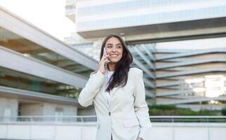 A businesswoman in a white blazer smiles while talking on a cell phone outside of a modern building. The bright light and sleek design of the building emphasize the professional nature of the scene. photo