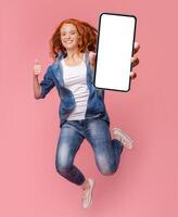 Cheerful girl redhead teenager jumping up, showing thumb up and newest smartphone with empty screen, enjoying new application for mobile phone. Pink studio background, creative image photo