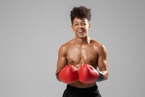 A young man boxer, displaying a confident smile, stands with his red boxing gloves raised. The studio setting features a neutral background, emphasizing his physique and dedication to the sport photo