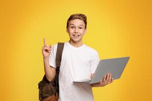 Cheerful boy holding laptop excited with idea or question, pointing finger up, number one, orange studio background with empty space photo