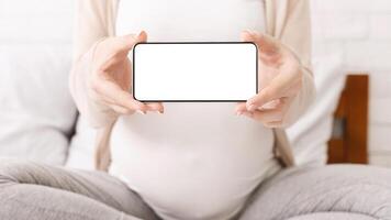A pregnant woman sits cross-legged on a bed in a bright room. She is holding a smartphone with a blank, white screen, possibly for a maternity or prenatal-related purpose, cropped photo