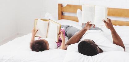 Black father and his daughter are lying in bed together, each reading their own book. Both are smiling and appear to be enjoying their time together. The setting is a cozy bedroom photo