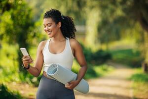A smiling woman with curly hair is walking in a park while holding a rolled-up yoga mat in her right hand and using her smartphone with her left hand, copy space photo
