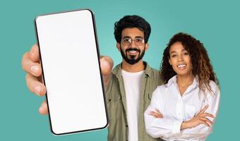 Indian man is holding up a smartphone with a blank white screen. The phone is in the foreground, and a man and woman are behind it, both smiling at the camera. The background is a solid teal color. photo