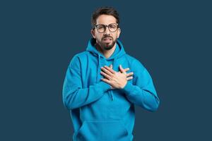 A young man in a blue hoodie stands with his hands on his chest, displaying a deep expression of emotion. The dark background highlights his thoughtful demeanor photo