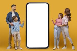 A smiling father, mother, daughter, and son stand together beside an oversized smartphone with a blank screen, possibly interacting with a digital application or advertisement photo