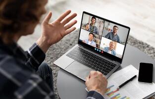 A man sits at a table, participating in a conference on his laptop. He waves his hand to the camera, engaging with the four other individuals in the call photo
