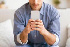 A man sits on a couch, holding a smartphone in his hands. He is looking down at the screen, focusing on his phone. photo