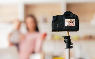In a bright, modern kitchen, a woman records a cooking tutorial using a professional camera set on a tripod. She appears to be explaining a recipe, with the camera capturing her in focus. photo